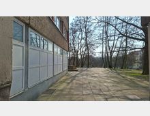 Gepflasterter Gehweg neben einem mehrstöckigen Wohngebäude mit großen verglasten Balkon- oder Loggiaelementen an der Fassade. Rechts stehen kahle Bäume auf einer Grünfläche, im Hintergrund sind weitere Wohnhäuser zu sehen.