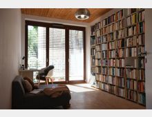 Wohnzimmer oder Arbeitszimmer mit bodentiefen Fenstern, die mit Jalousien ausgestattet sind und viel Tageslicht hereinlassen. Auf der rechten Seite steht ein großes, wandfüllendes Bücherregal, links ein Sofa und ein kleiner Schreibtisch mit einem Stuhl.
