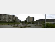 Panorama einer Wohnanlage mit mehreren mehrstöckigen, grauen Plattenbauten. Im Vordergrund befindet sich ein kleiner Springbrunnen mit umliegender gepflasterter Fläche, die von vereinzeltem Grün und Bäumen gesäumt ist.