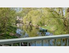 Ein kleiner Fluss mit klarer Wasseroberfläche, die die umstehenden grünen Bäume und Sträucher spiegelt, im Vordergrund ein grünes, vertäutes Boot und eine Holzbank am Ufer, im Hintergrund ein Gebäude und eine Brücke.