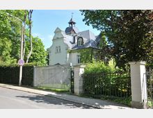 Historische Villa mit heller Fassade, verziertem Giebel und dunkelgrauem Schieferdach, umgeben von üppigem Grün und einer Metallzaunanlage mit Einfahrtstor. Die Straße im Vordergrund ist von Bäumen gesäumt, und ein Verkehrsschild weist ein Halteverbot aus.