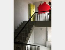 Treppenhaus mit weißen Wänden und einer Metallgeländer-Treppe. Im oberen Stockwerk sind eine gelbe Tür und eine rote, asymmetrische Wanddekoration zu sehen.