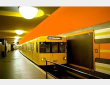 Eine U-Bahn-Station mit einem gelben U-Bahn-Wagen der Linie U7, der am Bahnsteig hält. Die Wände sind mit Mosaikfliesen in Gelb- und Orangetönen gestaltet, und die Decke ist mit runden, weißen Leuchten beleuchtet.