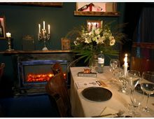 Ein stilvoll gedeckter Tisch in einem Restaurant mit Kerzenlicht, einem Blumenarrangement und einem "Reserviert"-Schild. Im Hintergrund befinden sich ein Kamin mit künstlichem Feuer, ein Bücherregal und dekorative Elemente an der dunkelgrünen Wand.