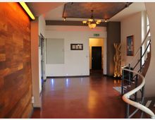 Ein Innenbereich mit modernem Design, bestehend aus einer rot-braunen Bodenfläche, einer Holzwand auf der linken Seite und einer geschwungenen Metalltreppe rechts. An der Decke befindet sich ein Kronleuchter, und der Raum ist mit indirekten Lichtelementen und Holzdekorationen gestaltet.