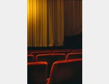 Innenraum eines Theaters oder Kinos mit leeren roten Stuhlreihen im Vordergrund und einem geschlossenen, gut beleuchteten goldfarbenen Vorhang auf der Bühne im Hintergrund.