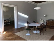 Großzügiger Wohnraum mit hellgrauen Wänden und Fischgrätparkettboden. Im Vordergrund steht ein weißer runder Tisch mit Holzstühlen auf einem Teppich, im Hintergrund ein weiteres Zimmer mit Sofa und Wandkunst, das durch eine breite Türöffnung sichtbar ist.