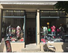 Fassade eines Designer-Outlets mit großen Schaufenstern, in denen Schaufensterpuppen mit Kleidung, Taschen und Accessoires ausgestellt sind. Auf der Glastür und den Fenstern sind Werbetexte aufgemalt, die Rabatte ankündigen; vor dem Eingang steht ein kleines Tisch- und Stuhl-Set.