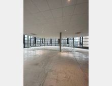 Leerer, moderner Innenraum mit Betonboden, Deckenplatten aus quadratischen Paneelen und großen, bodentiefen Fenstern, die einen Blick auf umliegende Gebäude bieten. Eine einzelne Säule steht mittig im Raum.