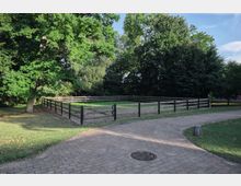 Grünfläche mit eingezäuntem Rechteckbereich, umgeben von hohen Bäumen, und einem gepflasterten Weg im Vordergrund. Der Zaun besteht aus Holz mit schwarzen Pfosten, und die Szene ist von einer natürlichen Parkumgebung geprägt.