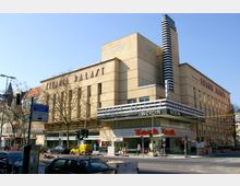 Außenansicht eines historischen Kinos namens "Titania Palast" mit beigem Fassadenputz und markantem rechteckigen Aufsatz links. Vor dem Gebäude befinden sich Geschäfte, Parkplätze, Straßenschilder und eine Ampelanlage.