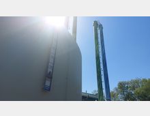 Großer zylindrischer Industriebau mit Meßskala an der Vorderseite, daneben bunt bemalte, hohe Schornsteine. Im Hintergrund ein klarer blauer Himmel und Bäume.