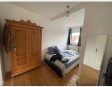 Schlafzimmer mit Holzboden, einem rustikalen Kleiderschrank aus Holz, einem Bett mit gemusterter Bettwäsche, einem Wandspiegel und einem Fenster mit blauen Vorhängen. Im Hintergrund ist eine weiße Wand und eine Zimmertür zu sehen.