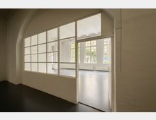 Heller, leerer Innenraum mit großer, bogenförmiger Glas-Trennwand und breiter Durchgangsöffnung zwischen zwei Räumen. Im Hintergrund hohe Sprossenfenster, weiße Wände, glatter Boden und Heizkörper entlang der Fensterfront.