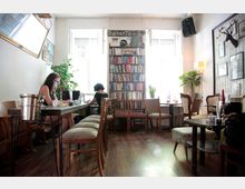 Gemütliches Café mit Holzböden und Bücherecke, in dem zwei Personen an Laptops arbeiten. Die Einrichtung umfasst Holztische, gepolsterte Stühle und Dekorationselemente wie Topfpflanzen, Bücherregale und eine Wand mit einem Hirschkopf-Dekor.