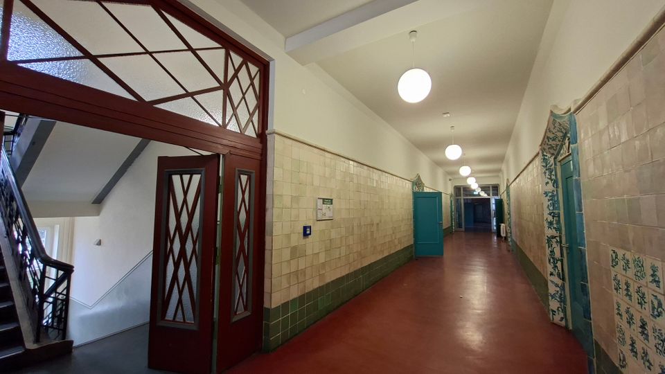 Langer Innenflur eines älteren Gebäudes mit roten Bodenbelag, halbhoch gefliesten Wänden und runden Deckenleuchten. Links befindet sich ein Treppenhaus mit Holz-Glas-Tür und Oberlicht, entlang des Flurs stehen mehrere türkisfarbene Türen mit dekorativen Fliesenrahmen.