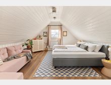 Gemütliches Schlafzimmer unter dem Dach mit weißer Holzverkleidung, einem Doppelbett, einer rosa Couch, einem geometrischen Teppich und minimalistischem Dekor. Ein Fenster mit beigen Vorhängen lässt Tageslicht herein, während Pflanzen und Bilder das Zimmer schmücken.