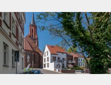 Straßenszene mit einer roten Backsteinkirche im neugotischen Stil im Hintergrund, flankiert von modernen Wohnhäusern mit weißen und roten Fassaden. Im Vordergrund sind ein Baum, parkende Autos und gepflasterte Straßen zu sehen.