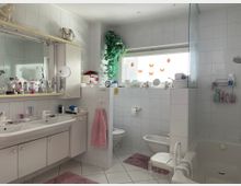 Ein helles Badezimmer mit weiß gefliesten Wänden und Boden, ausgestattet mit Doppelwaschbecken, eingelassener Badewanne mit Duschbereich, Toilette und Bidet. Das Fenster ist mit Schmetterlings- und Marienkäfer-Aufklebern dekoriert, und auf dem Waschtisch stehen viele Pflegeprodukte.