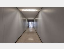 Langer, schlichter Flur in einem Gebäude mit weißen Wänden und einer abgehängten Decke mit Rasterleuchten. Entlang des Flurs befinden sich Türen, ein Feuerlöscher steht an der Wand, und am Ende des Gangs ist ein Durchgang mit Tageslicht erkennbar.