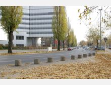 Moderne mehrstöckige Bürogebäude an einer breiten, von Bäumen gesäumten Straße mit parkenden Autos und Herbstlaub auf dem Gehweg. Im Vordergrund stehen mehrere runde Betonpoller entlang des Gehwegs.