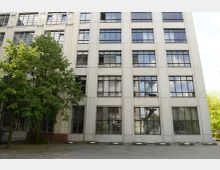 Fassade eines mehrstöckigen Gebäudes mit großen Fenstern und heller, teils verklinkerter Außenwand. Vor dem Gebäude stehen zwei grüne Bäume, und der Boden ist ein gepflasterter Parkplatz mit markierten Stellplätzen.