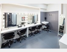 Schminkraum mit einem langen weißen Schminktisch, beleuchteten Spiegeln, vier schwarzen Drehstühlen und Kosmetikartikeln. Rechts befinden sich ein Waschbecken mit Spiegel und eine graue Fliesenwand.