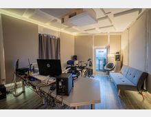 Musikstudio mit schallisolierten Wänden und Deckenplatten, ausgestattet mit einem Schreibtisch voller Technik, einem elektronischen Schlagzeug, Lautsprechern, einem grauen Sofa und einer Glastür mit der Aufschrift "Staff Only". Der Raum ist modern eingerichtet und mit Holzfußboden ausgelegt.