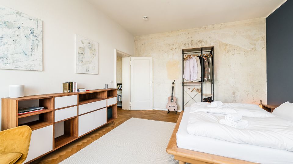 Helles Schlafzimmer mit Holzbett und weißer Bettwäsche auf Fischgrätparkett, dazu ein langer Holz-Sideboard mit offenen Fächern und abstrakten Bildern an der Wand. An der Rückwand stehen ein offenes Kleiderregal und eine Gitarre vor einer teils roh wirkenden, hellen Wandfläche.