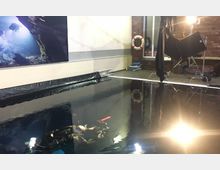 Indoor diving training pool with dark water, bordered by black plastic sheeting and surrounded by brick and white walls. Studio lighting equipment and a wall-mounted life ring are visible around the pool’s edge.