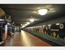 U-Bahn-Station mit geschwungener Decke, beleuchtet durch runde Deckenleuchten. Die Wände sind mit bunten Mosaiken in Blumenmotiven dekoriert, und der Schriftzug "Paulsternstraße" ist an einer der Wände angebracht.