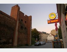 Backsteinmauer eines historischen Gebäudes mit gotischen Fensteröffnungen auf der linken Seite, daneben eine Straßenszene mit parkenden Autos, einer Restaurant-Leuchtreklame und weiteren Häuserfassaden im Hintergrund. Ein Aufsteller mit der Aufschrift „Echte Prager Küche“ steht am Gehweg.