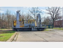 Gleiskran auf einer Schienenanlage mit Betonboden, umgeben von kahlen Bäumen und einem roten Backsteingebäude im Hintergrund.