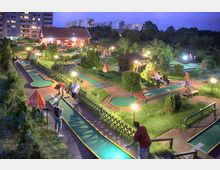Miniaturgolfplatz bei Dämmerung mit beleuchteten Bahnen, umgeben von Rasenflächen, Bäumen und kleinen Sträuchern; im Hintergrund ein Gebäude mit rotem Dach und moderne Wohngebäude. Spielende Personen und Sitzplätze mit Sonnenschirmen sind sichtbar.