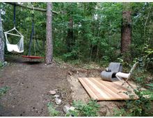 Waldszene mit zwei Schaukeln, die an Bäumen befestigt sind, und einem kleinen Holzpodest mit einem modernen Stuhl und einer weißen Gartenstuhl daneben. Umgeben von dichtem, grünem Laub und hohen Bäumen.