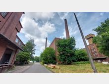 Industriegelände mit Backsteingebäuden und hohen Schornsteinen, umgeben von Bäumen und einer asphaltierten Straße. Im Hintergrund sind ein Wachturm und weitere industrielle Strukturen sichtbar, während Holzstapel am Straßenrand liegen.