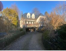 Ein zweistöckiges Wohnhaus mit heller Fassade und grauem Schieferdach, umgeben von dichter Vegetation und kahlen Bäumen. Im Vordergrund führt eine gepflasterte Einfahrt zu einer offenen Garage unterhalb des Hauses.