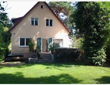 Ein zweistöckiges Einfamilienhaus mit beiger Fassade und roten Dachschindeln, umgeben von einem gepflegten Garten mit Rasen und Bäumen. Auf der Rückseite des Hauses führt eine Treppe von einer Terrasse mit Pflanzkübeln in den Garten.