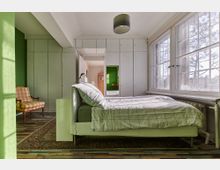 Schlafzimmer mit grün gestrichenen Wänden, einem Bett mit kariertem Bettzeug und einer großen Fensterfront mit Blick nach draußen. Im Raum befinden sich zudem ein Sessel, ein bunter Holzboden, Einbauschränke und ein Spiegel an der Wand.