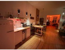 Gemütliche Wohnküche mit Holzboden und warmem Licht; links eine Küchenzeile mit Holzarbeitsplatte, weißem Smeg-Kühlschrank und Hängelampen, dahinter ein Essbereich mit Holztisch und Stühlen. Im Hintergrund ein offener Wohnraum mit großen Fenstern.