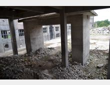 Verlassenes Industriegebäude mit Betonstützen und zerbrochenen Fenstern, umgeben von Schutt- und Trümmerhaufen. Im Hintergrund sind freiliegende Wände und eine offene Fläche mit mehr Trümmern sowie vereinzelten Bäumen sichtbar.