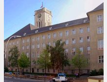 Großes Verwaltungsgebäude mit hellem Putz und zahlreichen Fenstern, darüber ein Uhrturm mit einer Fahne auf der Spitze. Im Vordergrund stehen Bäume, parkende Autos und eine Straße.