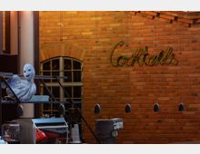 Eine Bar mit einer Backsteinwand im Hintergrund, auf der der Schriftzug „Cocktails“ in gelber Neon-Leuchtschrift angebracht ist. Im Vordergrund befinden sich eine Espressomaschine, eine Figur in Form eines Geistes auf einem Regal und Lampen, die von der Decke hängen.