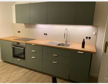 Moderne Küche mit grünen Schränken, einer Arbeitsplatte aus Holz, einem integriertem Backofen und Glaskeramikkochfeld. Darüber befinden sich Hängeschränke, eine Edelstahlspüle mit hohem Wasserhahn, und eine helle Fliesenrückwand.