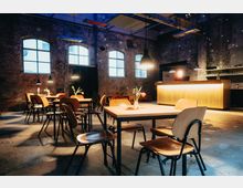 Industrie-inspirierter Caféraum mit Backsteinwänden, großen Fenstern und sichtbarer Decke. Holzstühle und -tische mit Vasen und einzelnen Blumen, dazu warme Beleuchtung und eine beleuchtete Theke im Hintergrund.