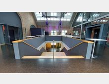 Innenraum eines modernen Veranstaltungsgebäudes mit Backsteinwänden, großen Fenstern und einem offenen Treppenbereich in der Mitte. Die Treppen führen hinunter zu einem unteren Bereich, während Oberlichter in der Decke Tageslicht einlassen.