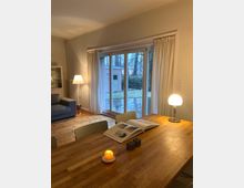 Ein gemütlicher Wohnraum mit Holzboden, einer Holztafel mit einem aufgeschlagenen Buch, einer brennenden Kerze und einer Tischlampe. Im Hintergrund befinden sich eine bodentiefe Fenster­schiebetür mit Vorhängen und ein Blick auf einen regennassen Garten.