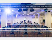 Ein moderner Konferenzraum mit langen weißen Tischen und schwarzen Stühlen, die in Reihen aufgestellt sind. Im Hintergrund befinden sich zwei Kamerastative, Werbewände mit dem Schriftzug "Mercure" und breite Vorhänge sowie Deckenbeleuchtung.