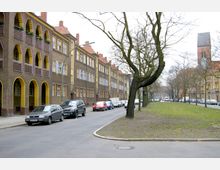 Straßenszene in einer Wohngegend mit mehrstöckigen Häusern im Stil der 1920er Jahre, gekennzeichnet durch separate Arkaden und gelb umrahmte Fenster. Entlang der Straße stehen parkende Autos, und auf dem Grünstreifen in der Straßenmitte sind vereinzelt kahle Bäume zu sehen; im Hintergrund ist der Turm einer Backsteinkirche erkennbar.
