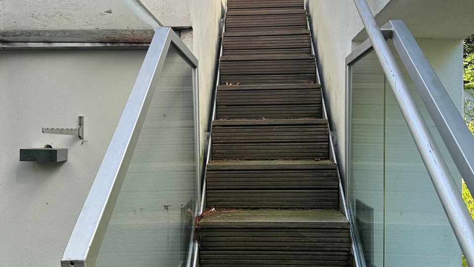 Außentreppe aus Holz zwischen zwei weißen Gebäudewänden, mit Glasgeländern und Metallhandlauf auf beiden Seiten. Oben führt die Treppe zu einer kleinen Plattform mit großer Satellitenschüssel vor blauem Himmel und Bäumen im Hintergrund.
